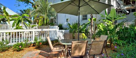 A patio with a table and chairs, a white picket fence, and various tropical plants.