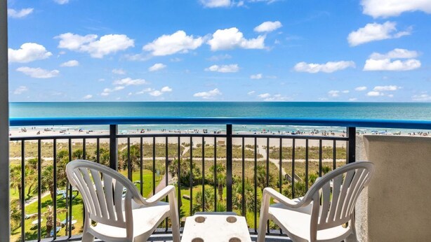 Un balcon avec deux chaises blanches et une petite table, donnant sur une plage, avec un ciel bleu dégagé et l’océan.