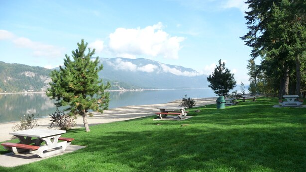 A lakeside picnic area with picnic tables and benches, surrounded by grass and trees, with mountains in the background. - 4 Bedroom Home in the Shuswap,  Close to Canoe Beach! (Canoe)