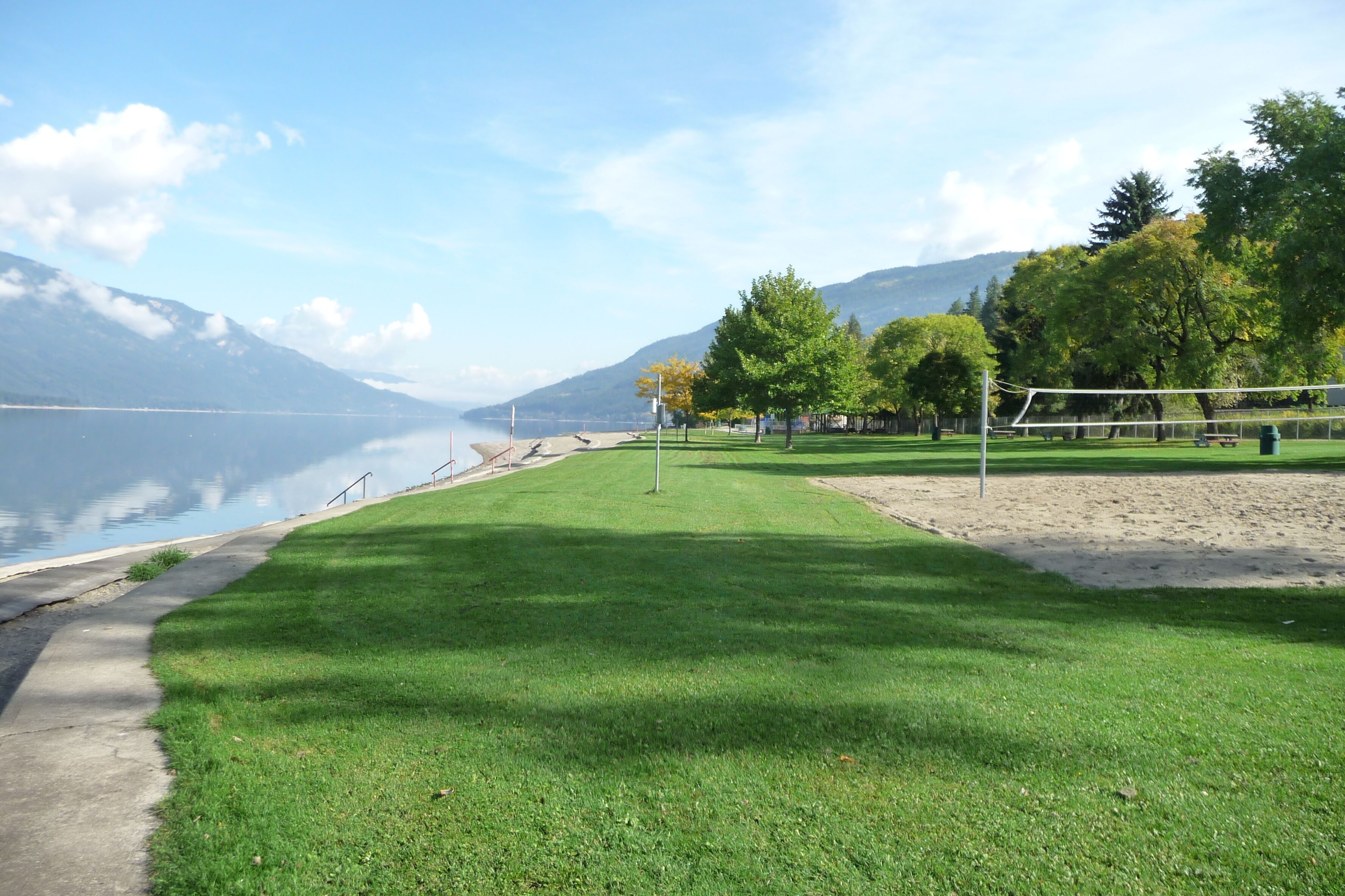 A lakeside park with a volleyball net, a concrete path, and trees.