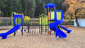 A playground with two blue slides, a climbing structure, and a netted area. - 4 Bedroom Home in the Shuswap,  Close to Canoe Beach! (Canoe)