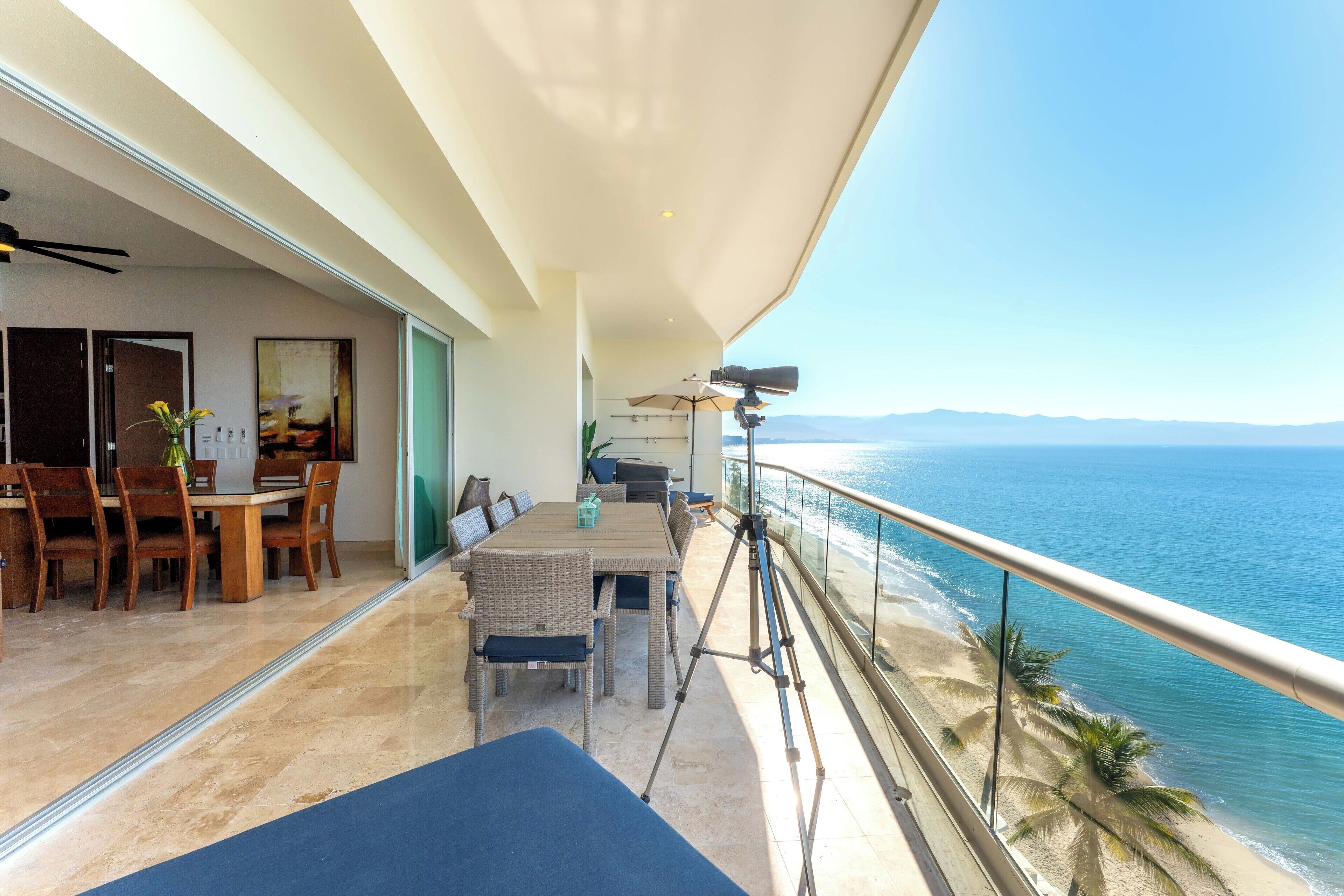 Un balcone con vista sull'oceano, un'area pranzo con sedie e tavolo, e una videocamera su un treppiede.