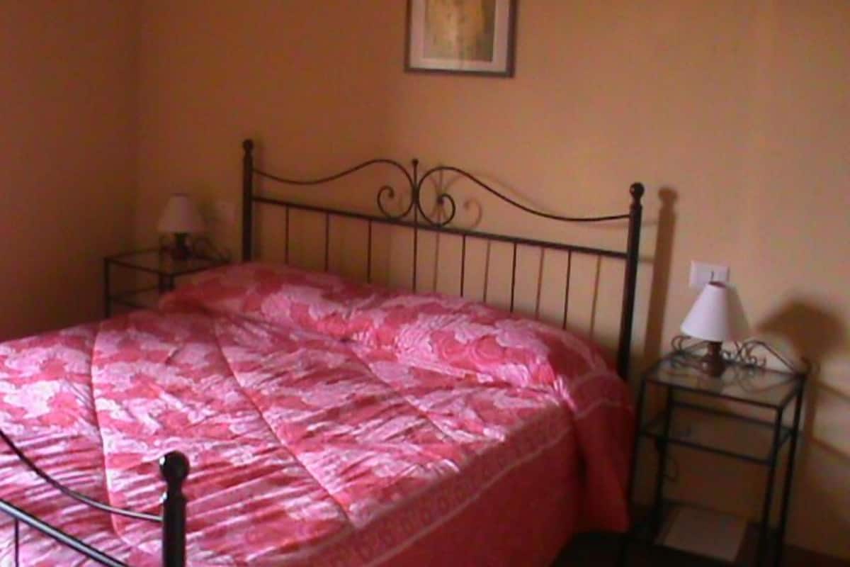 Un lit simple avec un cadre en métal et une couverture rose. Il y a une table de chevet avec une lampe, ainsi qu’une petite table sur laquelle se trouve une autre lampe.