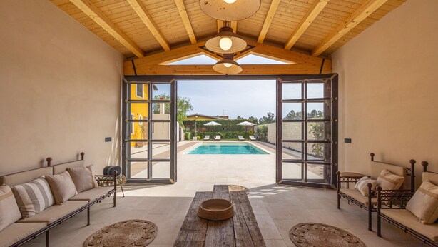 A spacious indoor area with a wooden ceiling, a pool visible through glass doors, and a seating area with a table and cushions. - Stylish Sicilian Villa with private pool near the Beach. (ispica)