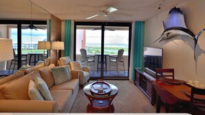 A hotel room with a sofa, a dining table, chairs, a ceiling fan, a mounted fish, and a TV.
