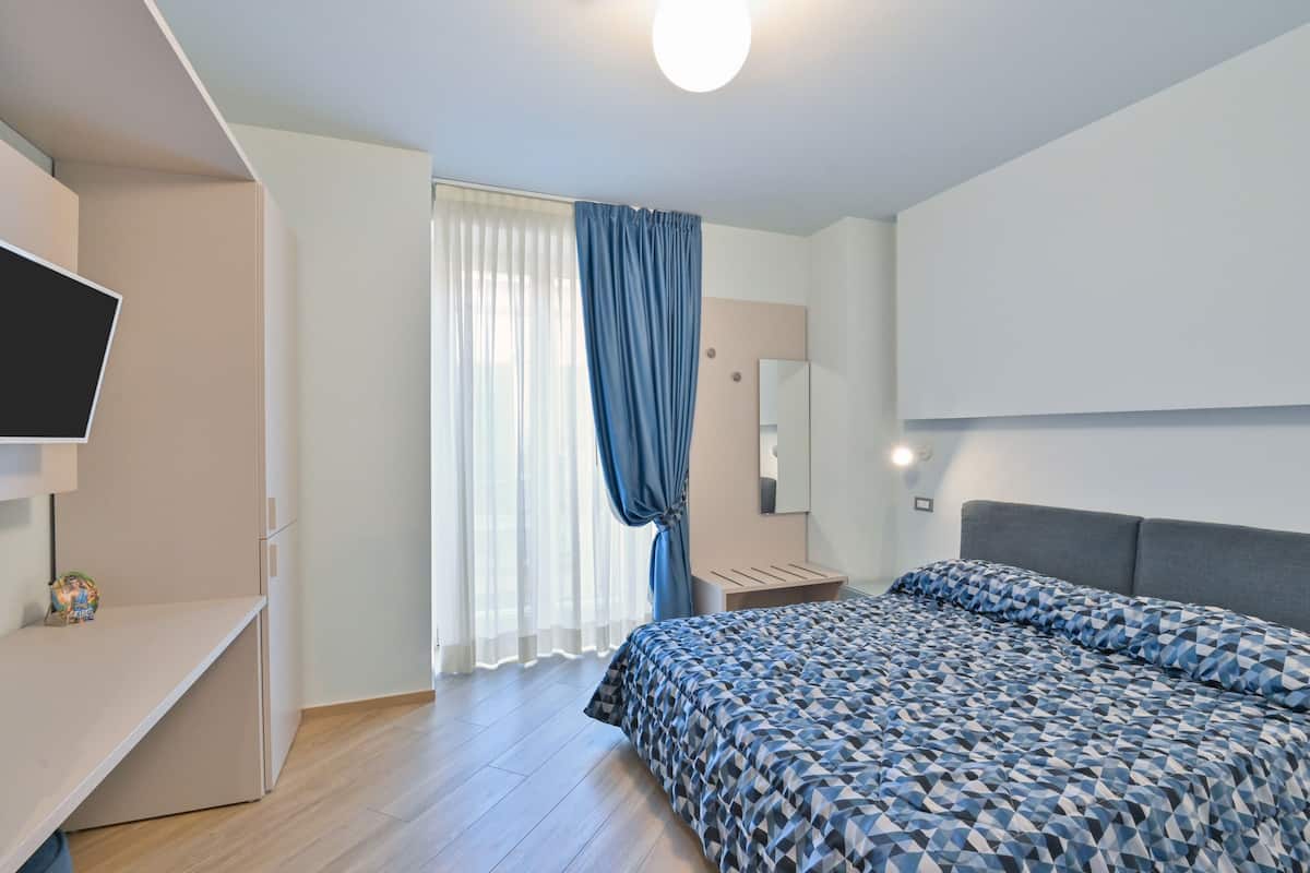 Ein modernes Schlafzimmer mit einem Bett, einem Fernseher, einem Schreibtisch, einem Spiegel und einem Fenster mit Vorhängen.