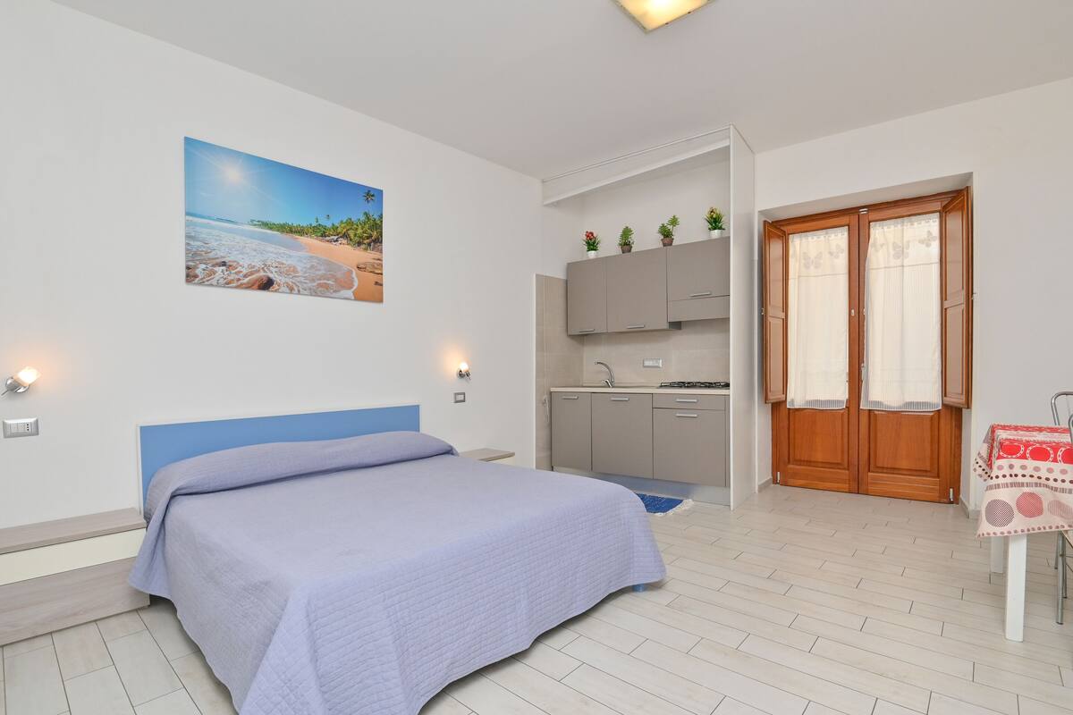 Ein Schlafzimmer mit Bett, einem Strandbild, einer Küchenzeile und einer Tür mit Vorhängen.
