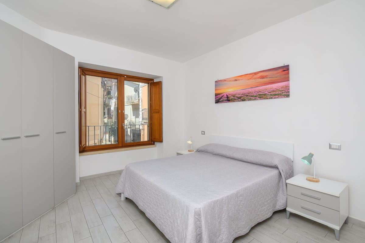 Ein Schlafzimmer mit einem Bett, einem Nachttisch, einer Lampe, einem Fenster und einem Gemälde an der Wand.