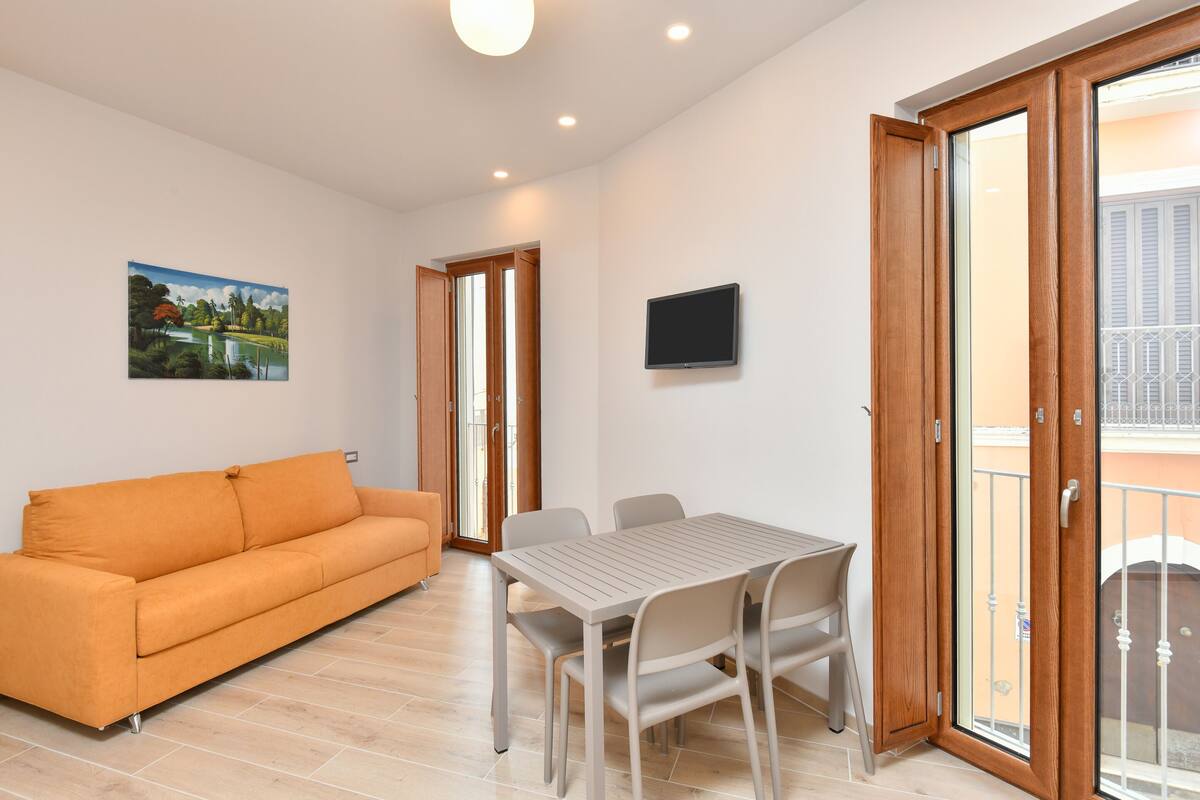 Ein modernes Wohnzimmer mit einer Couch, einem Esstisch und Stühlen. An der Wand ist ein Fernseher angebracht und eine Glasschiebetür führt in einen Außenbereich.