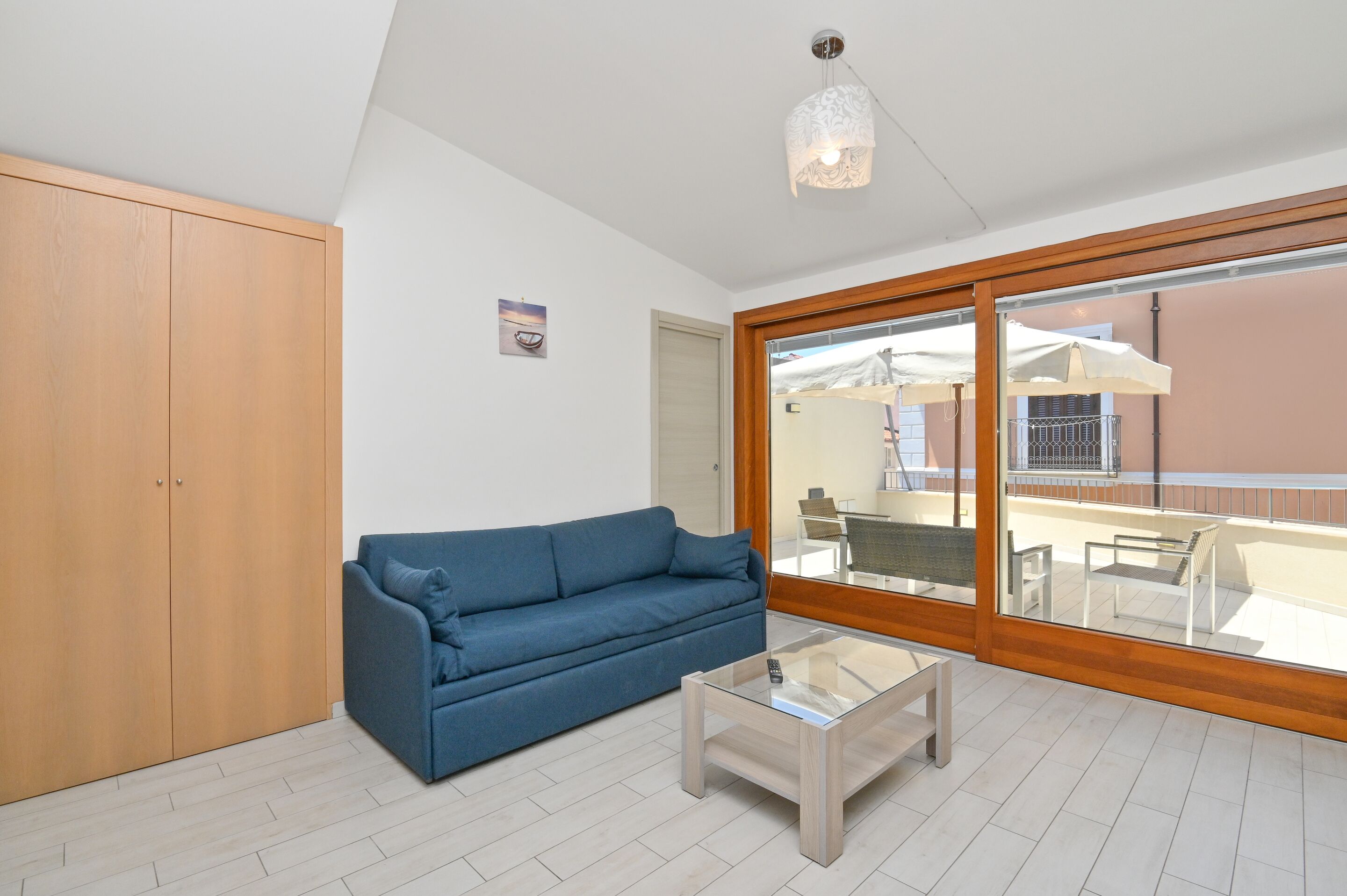 A room with a blue sofa, a glass coffee table, a wooden wardrobe, and a large sliding glass door leading to an outdoor area with a table and chairs.