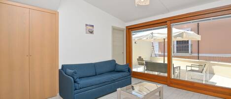 A room with a blue sofa, a glass coffee table, a wooden wardrobe, and a large sliding glass door leading to an outdoor area with a table and chairs.