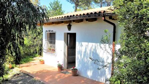 A small building with a tiled roof, a door, and a window with decorative bars. There are potted plants and a small garden area in front. - Casa Anselmi (Portoferraio)