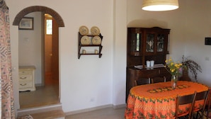 A dining area with a table covered in a floral tablecloth, wooden chairs, a hanging light fixture, and a wall-mounted shelf with decorative plates. - Casa Anselmi (Portoferraio)