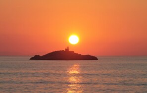 Un atardecer sobre un mar tranquilo con una pequeña isla y un faro a lo lejos.