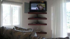 A living room with a television mounted on a wall, a sofa with striped cushions, and a small decorative item on a shelf. - Quaint Studio Cottage with private pool overlooking beautiful Coral Bay (Coral Bay)