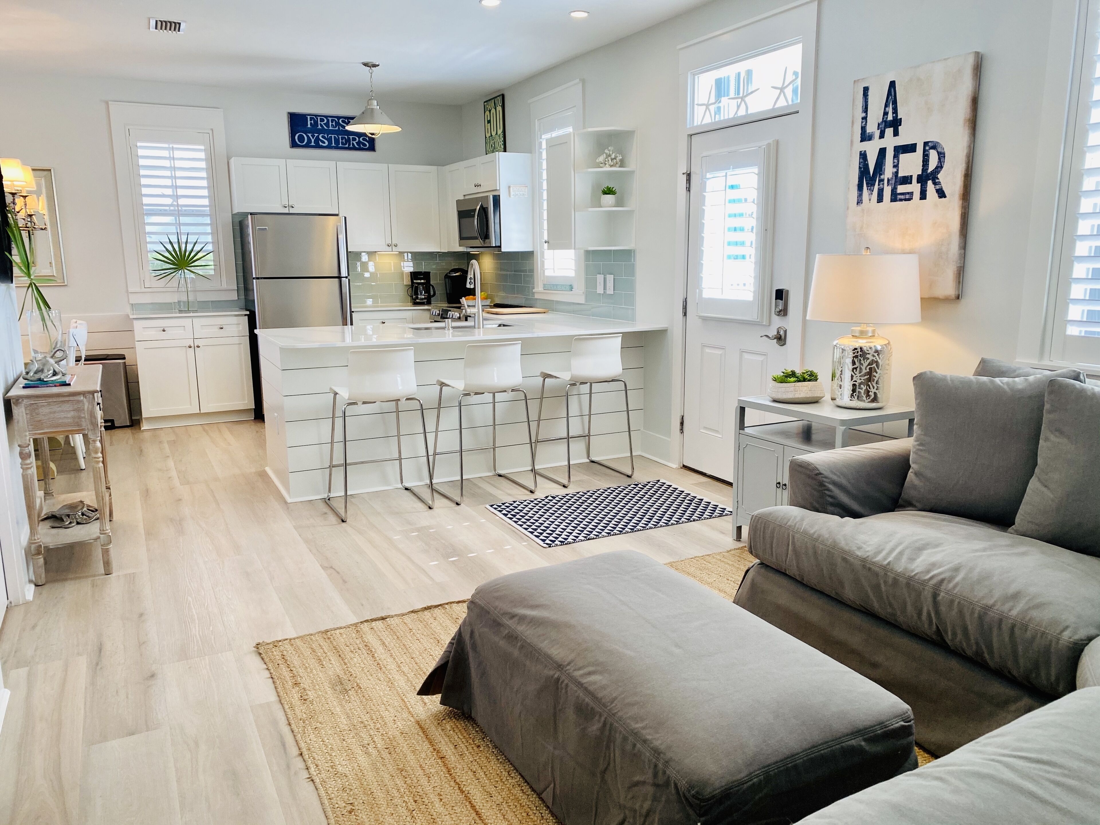 Cocina moderna con armarios blancos, refrigerador de acero inoxidable y una zona de bar con taburetes blancos. En la sala de estar, hay un sofá gris y un otoman gris.