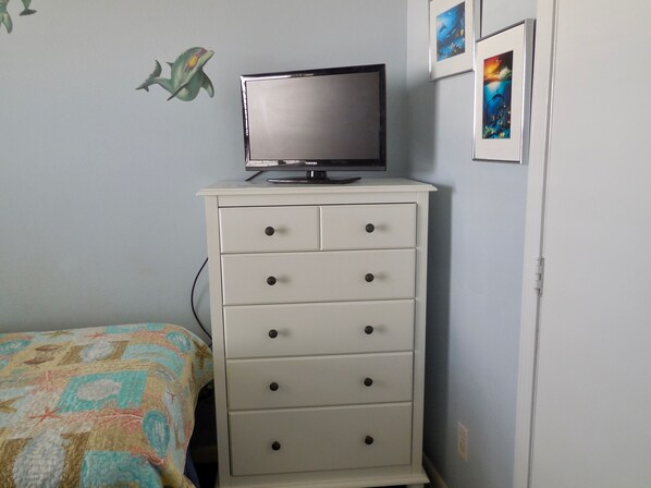 Een slaapkamer met een bed, een witte dressoir met lades, een televisie erbovenop en decoraties aan de muur.