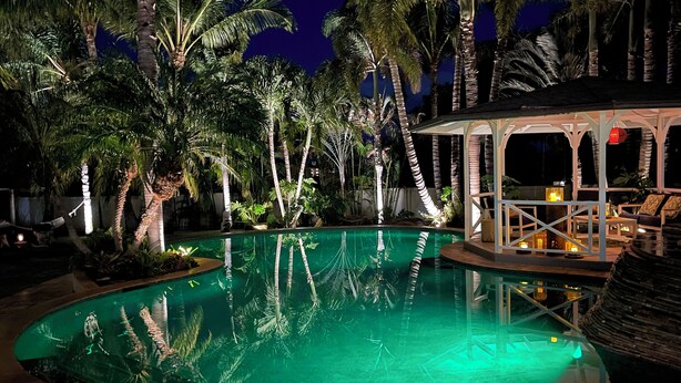 Un espace piscine nocturne avec une piscine à eau claire, entourée de palmiers et dotée d’un espace salon abrité.