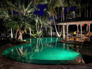 Un espace piscine nocturne avec une piscine à eau claire, entourée de palmiers et dotée d’un espace salon abrité.