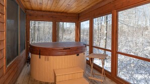 A wooden cabin with a hot tub, large windows, and a view of a snowy forest.
