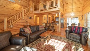 A spacious log cabin interior with a living area featuring leather sofas, a wooden coffee table, and a dining area with a kitchen in the background.