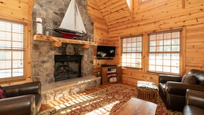 A cozy cabin interior with a stone fireplace, wooden walls, leather furniture, and a large window.
