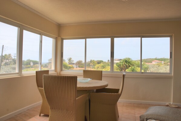 A room with a round table and four wicker chairs, large windows with a view of greenery and buildings, and a tiled floor.