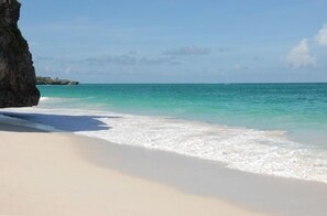 A beach with clear turquoise water, white sand, and a large rock formation on the left.