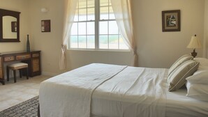 A bedroom with a large bed, a desk, and a chair. There is a window with curtains, a lamp, and a painting on the wall. - Your Relaxing Tropical Getaway Near Crane Resort - 3 bedroom (The Crane)