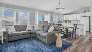 Eine moderne Küche mit weißen Schränken, ein Essbereich mit Holztisch und -stühlen sowie ein graues Ecksofa mit gemusterten Kissen.
