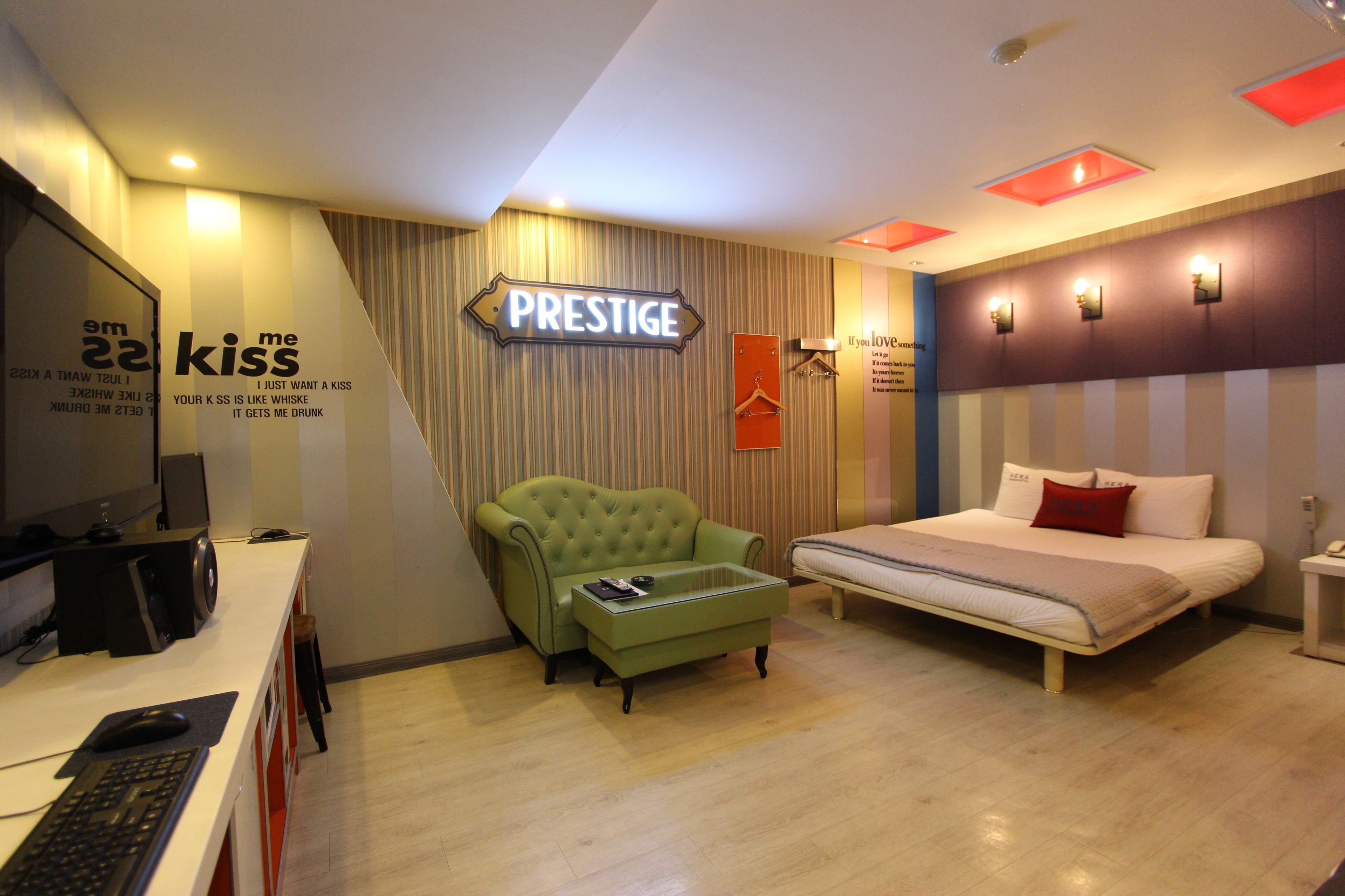 Ein modernes Hotelzimmer mit einem Bett, einem grünen Sessel, einem Schreibtisch mit Computer und einem Schild mit der Aufschrift 'PRESTIGE'.