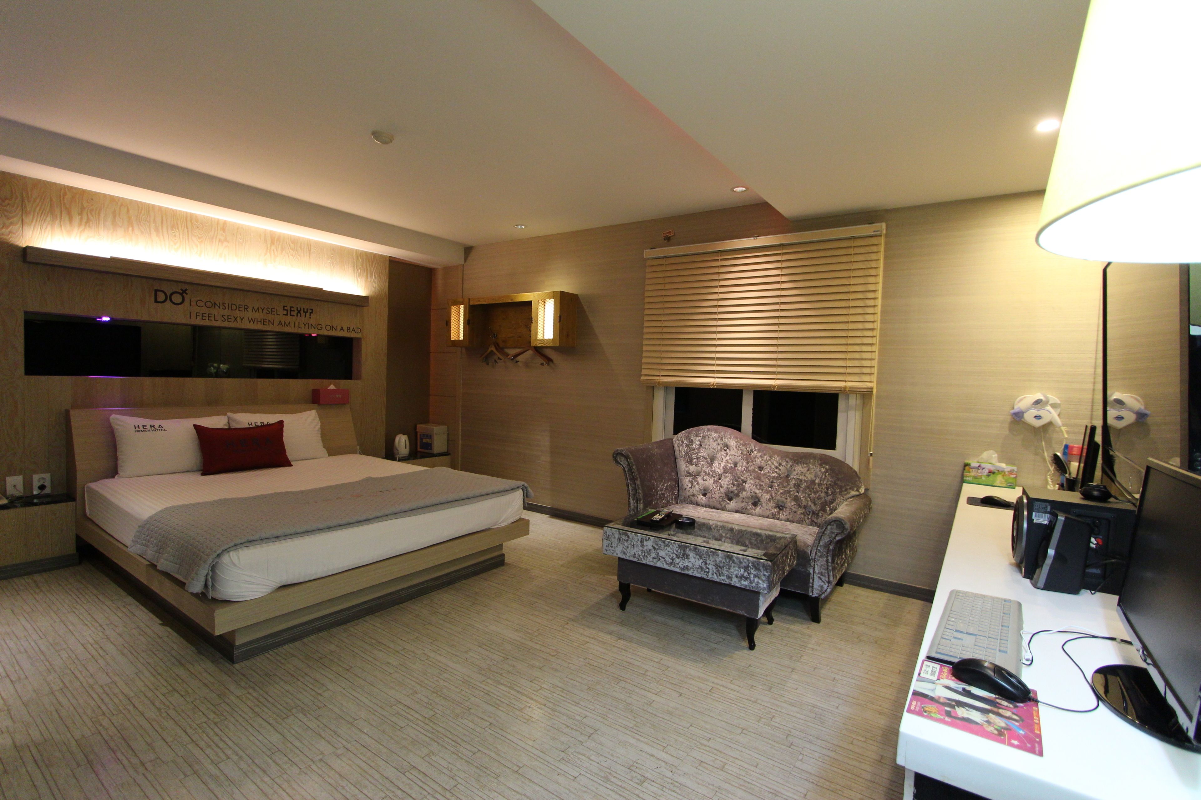 A hotel room with a bed, a sofa, a desk with a computer, and a window with blinds.
