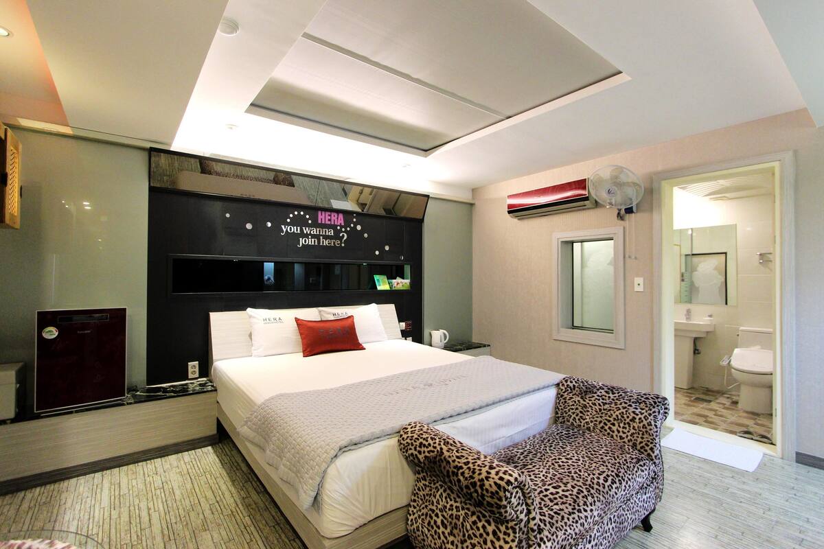 A modern hotel room with a large bed, a leopard print chair, a built-in TV, and a visible bathroom.