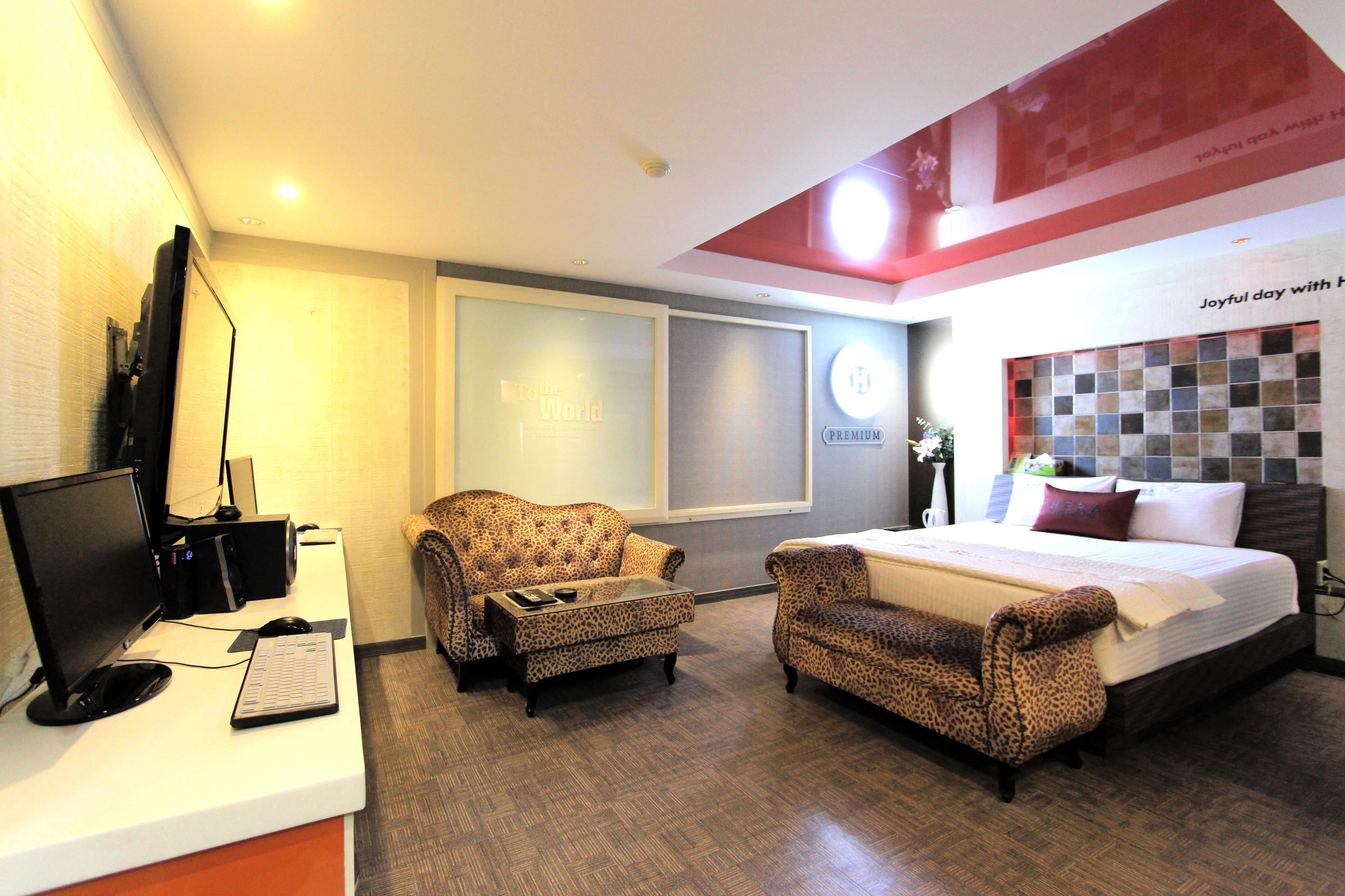 A hotel room with a bed, a desk with a computer, two armchairs, and a large window.
