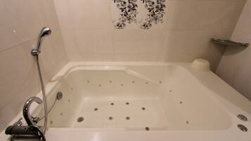 Una bañera blanca con ducha y mandos, un estante pequeño con dispensador de jabón, y un patrón de azulejos con motivos florales en la pared.