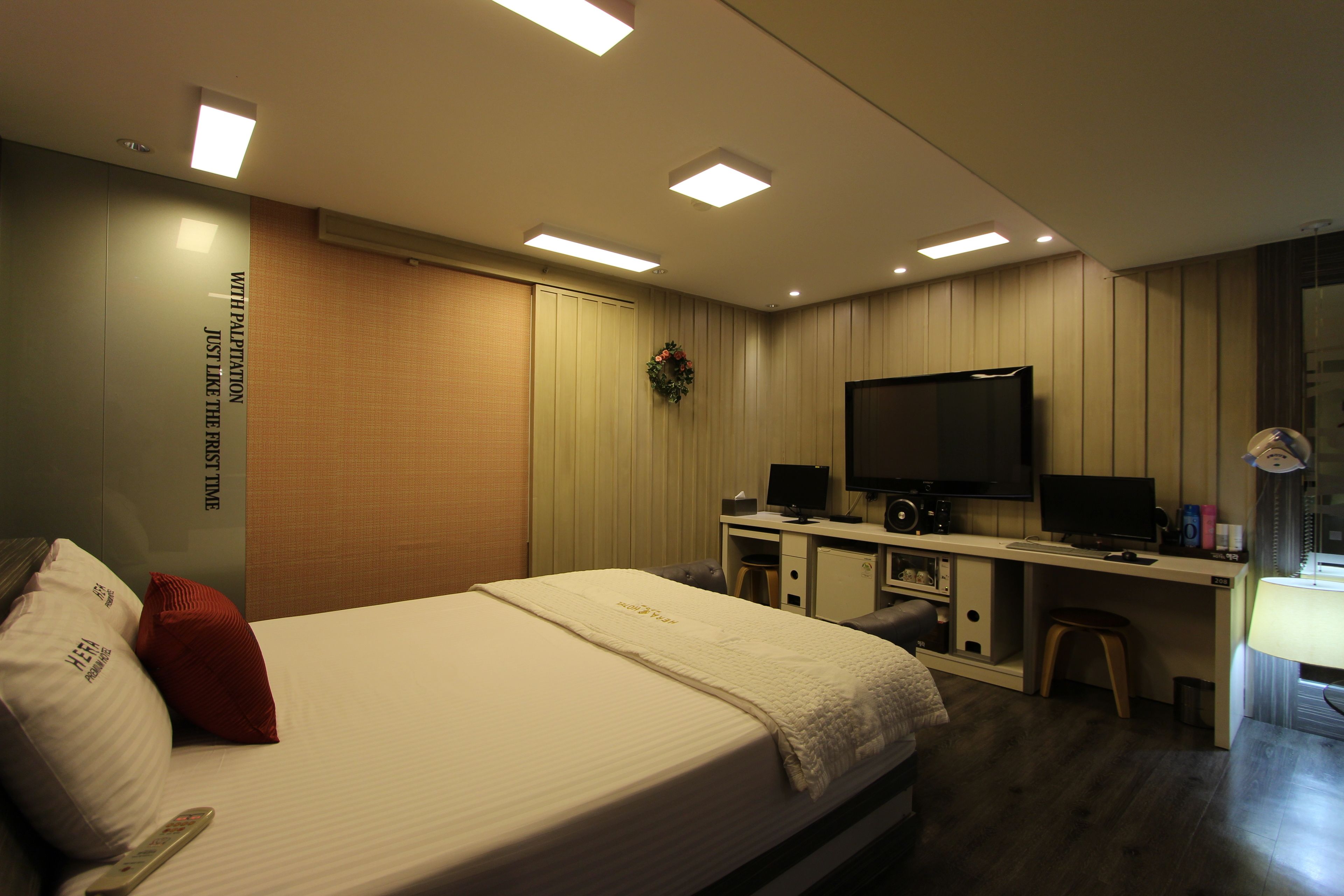 A hotel room with a bed, a desk with a computer, a TV, and a small stool.
