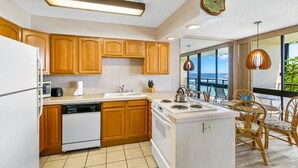 A modern kitchen with wooden cabinets, a white stove, and a view of the ocean through large windows. - A Beautiful Condo at the Waters Edge with a Great View (Koloa)