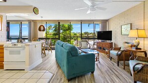 A spacious living area with a sofa, dining table, and chairs, a kitchen with a stove, and a large window offering a view of the outdoors. - A Beautiful Condo at the Waters Edge with a Great View (Koloa)