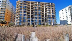 A multi-story residential building with balconies, a pathway in the foreground, and a clear blue sky.