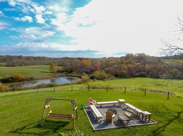 A picnic area with a swing set, benches, and a fire pit, surrounded by a grassy field and a pond, with trees in the background.