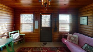 A log cabin interior with a wooden door, two windows with blinds, a green chair, a pink sofa, and a patterned rug.