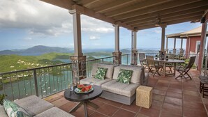 Eine überdachte Terrasse mit Gartenmöbeln, einem Tisch mit Wassermelone und Getränken sowie Blick auf einen See und Berge.