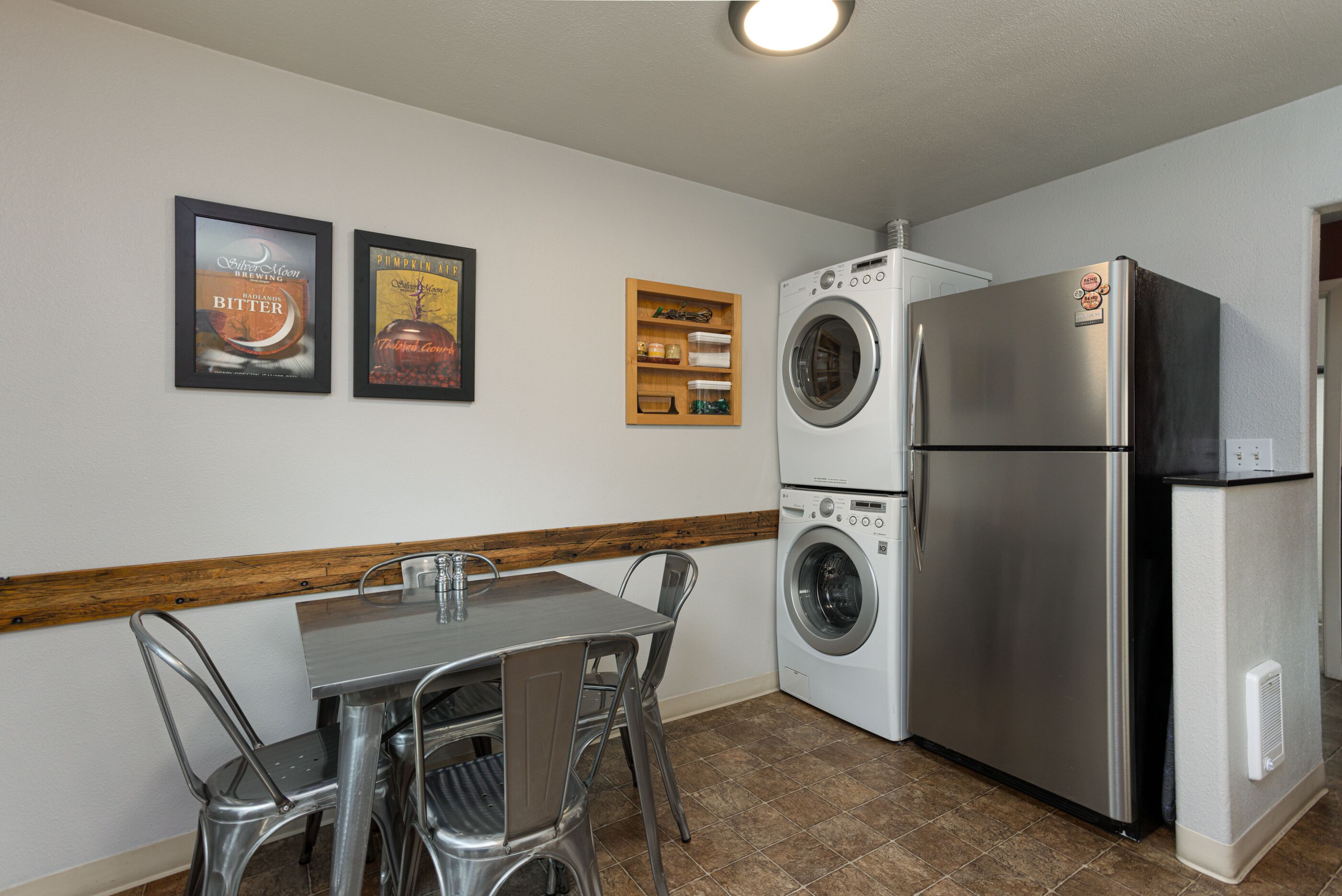 A compact kitchen with a stainless steel refrigerator, a double washer and dryer, a wooden shelf with jars, and a framed picture of a beverage.