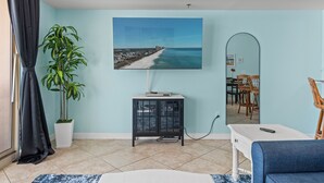 Una habitación con una televisión grande mostrando una escena de playa, una planta en maceta, un armario y un comedor con sillas y una mesa.