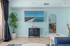 A room with a large TV displaying a beach scene, a potted plant, a cabinet, and a dining area with chairs and a table.