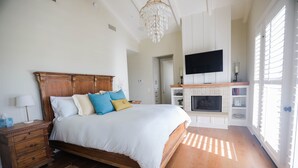A bedroom with a wooden bed, a nightstand, a TV, a fireplace, and a large window with blinds. - Playa Penthouse (Ocean Views) (Carpinteria)