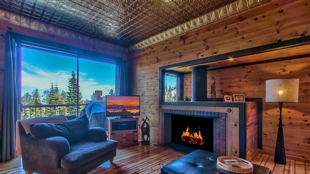 L’intérieur chaleureux d’un chalet avec une cheminée, un canapé, un téléviseur et une grande fenêtre donnant sur une forêt.