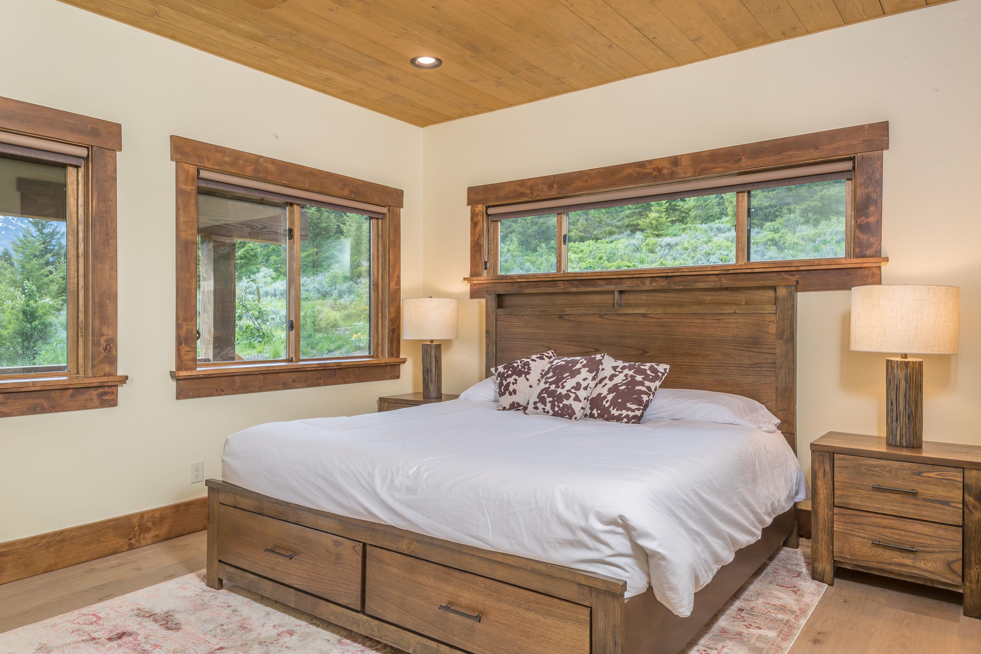 Une chambre à coucher avec un lit en bois, deux tables de chevet avec des lampes et de grandes fenêtres avec des cadres en bois.