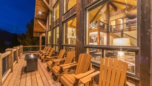 Une terrasse en bois, dotée de chaises Adirondack et d’un foyer, offre une vue sur un bâtiment avec de grandes fenêtres et un escalier.