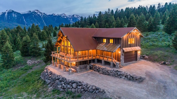 Une cabane en rondins, avec des fondations en pierre, une terrasse en bois et un grand garage. Elle est entourée d’un mur de pierre et se trouve dans une zone boisée, avec des montagnes en arrière-plan.